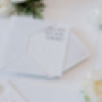 Embossed white invitation card partially in envelope with text "Will we see you there?" surrounded by white flowers and greenery.