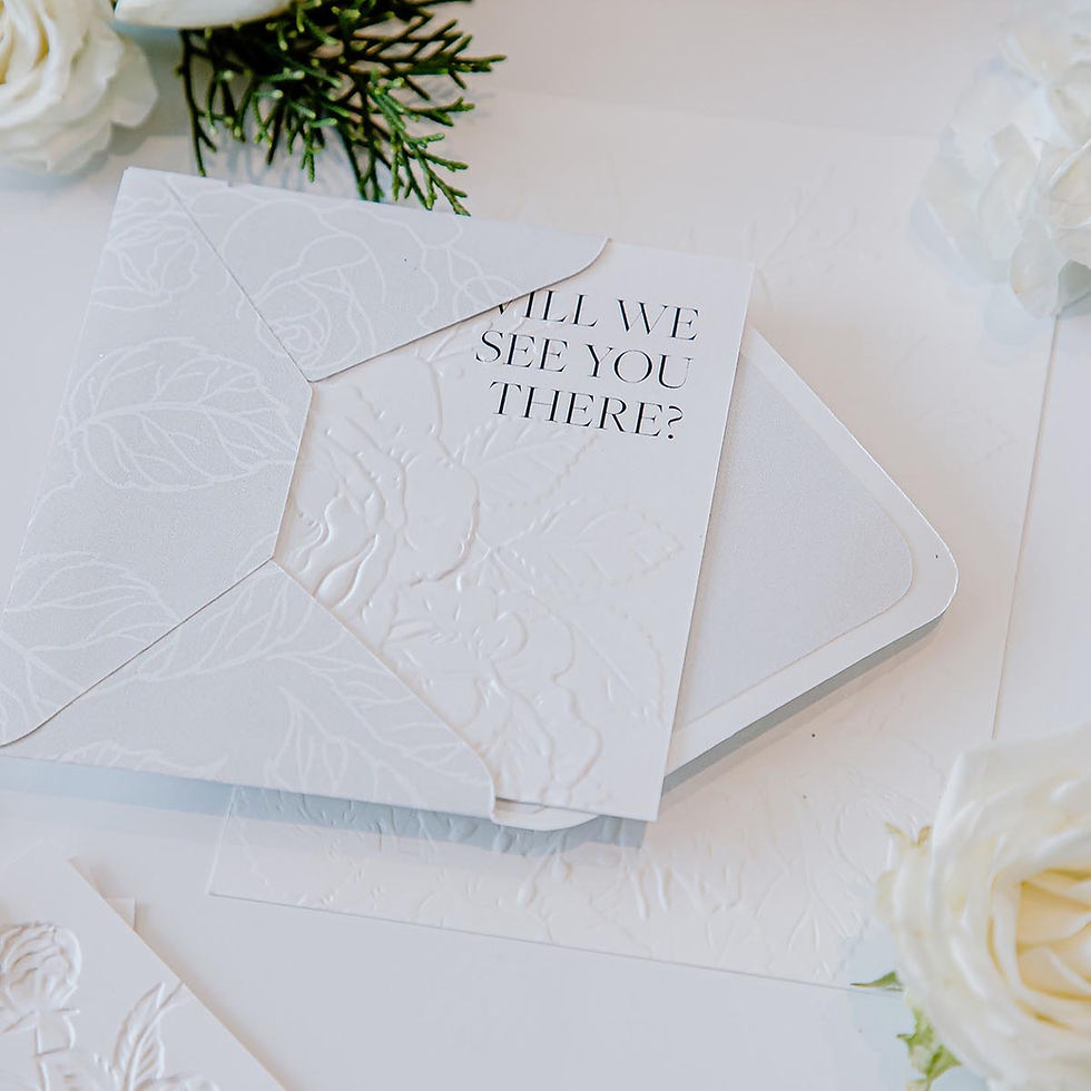 Embossed white invitation card partially in envelope with text "Will we see you there?" surrounded by white flowers and greenery.