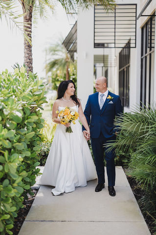 Bride and groom walking hand in hand along a garden path, bride holding yellow bouquet, both smiling at each other.