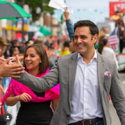Hernán Fratto, in gray blazer, high-fives crowd at lively outdoor event. Woman in pink scarf smiles. Colorful flags and signs in background.