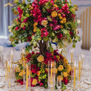 Tall Mediterranean wedding centerpiece with bougainvillea, yellow roses, greenery, and tapered candles on a gold-accented table.