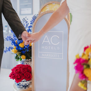 Bride and groom holding hands in front of AC Hotel welcome sign with Mediterranean-inspired florals.