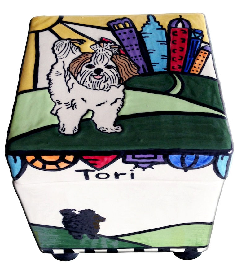 Thumbnail: Small white dog with bow on green hill, colorful city buildings and yellow sun in background – dog box urn for Tori