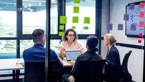 Team meeting in a glass conference room, four coworkers discuss plans around a laptop and colorful sticky notes.