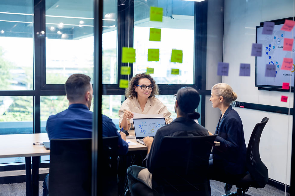 Team meeting in a glass conference room, four coworkers discuss plans around a laptop and colorful sticky notes.