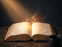 A glowing open book with magical particles rising from its pages, accompanied by a pair of reading glasses and a mug on a rustic wooden table.