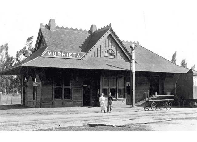 black and white photo of wooden building that used to be Murrieta's Train Depot with 2 people standing outside of it.