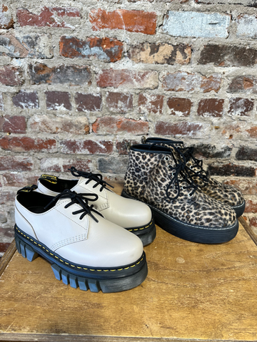 Two pairs of platform shoes, one white and one leopard print, displayed on wood surface with brick wall background.