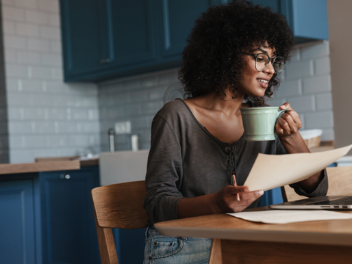 woman with glasses at kitchen table, working on laptop with green mug in hand
