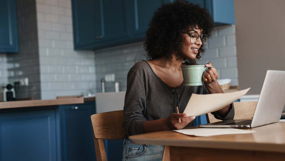 Woman with glasses sitting at kitchen table in front of laptop with green mug in hand
