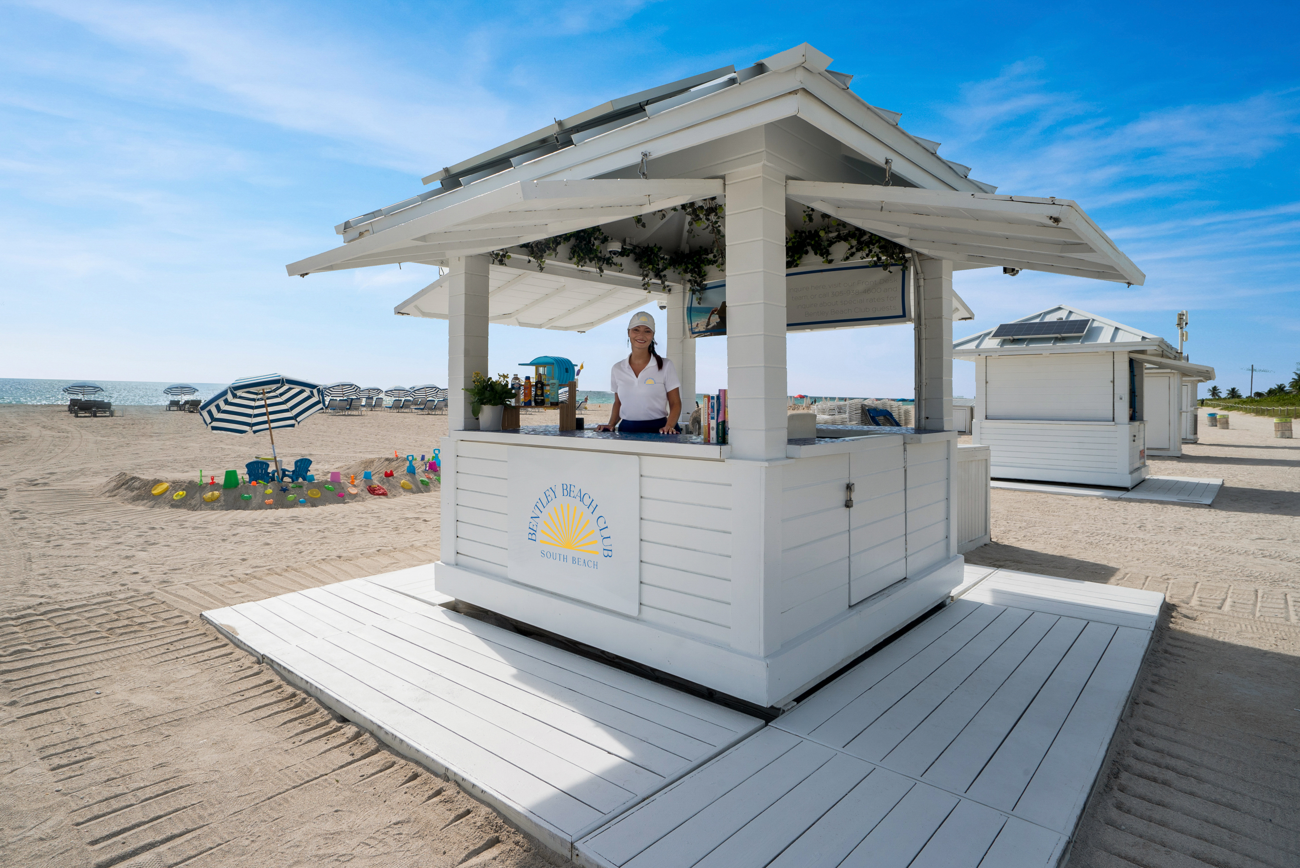A white beach hut with a woman standing inside on a sandy beach with a blue sky in the background.