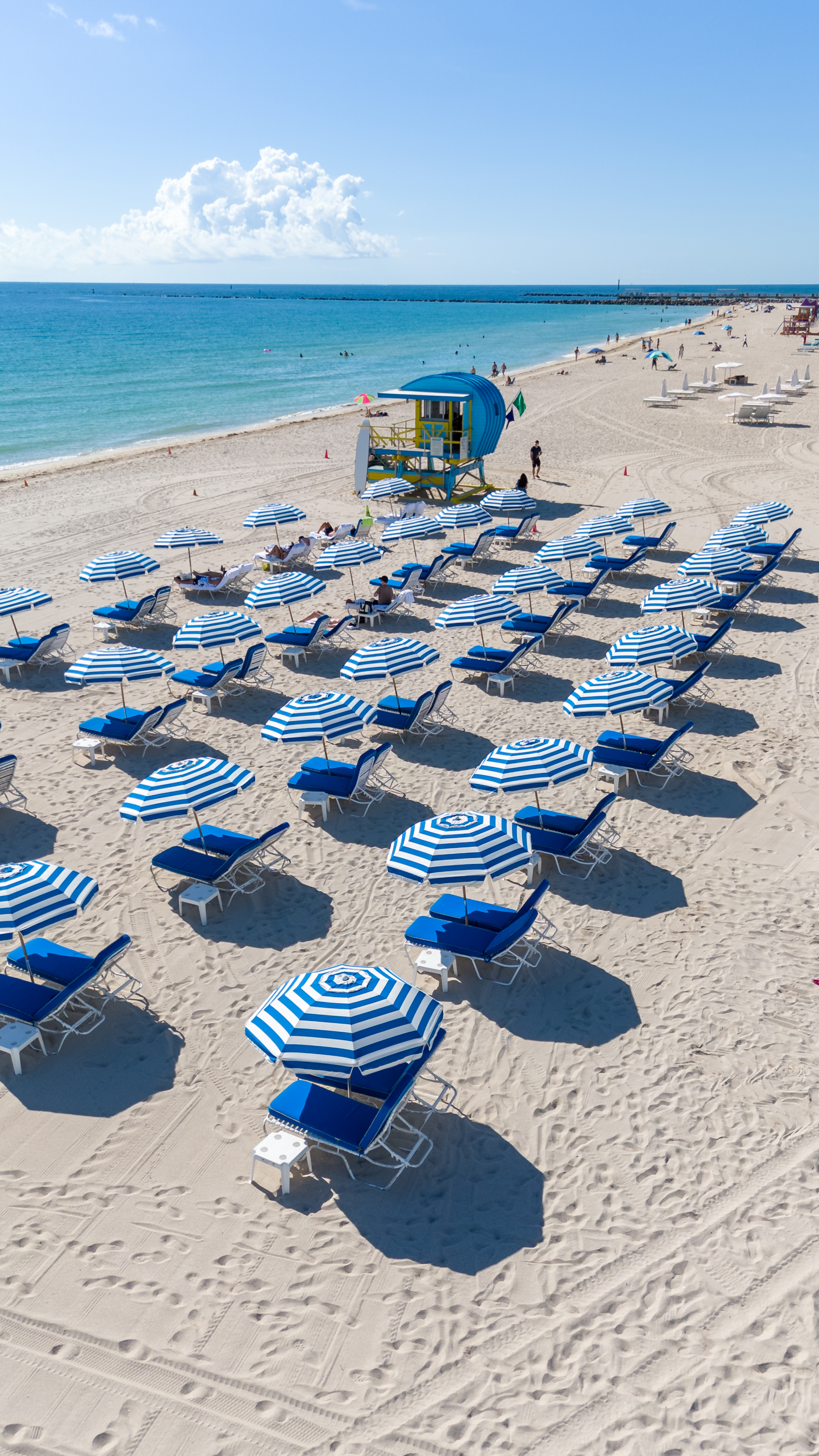 A beach scene with blue and white umbrellas and chairs on the sand, a lifeguard tower in the background, and the ocean in the distance.