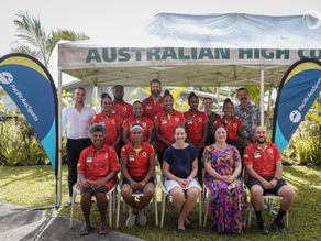 Group photo of the Vanuatu Women’s Cricket Team with staff and Australian High Commission representatives under a tent labeled “AUSTRALIAN HIGH COMMISSION.” Two PacificAus Sports banners are displayed on either side. The team members are wearing red uniforms, and the setting is an outdoor garden area.