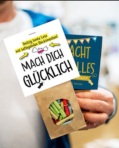 Auf dem Bild ist ein Losbeutel zu sehen. Im Vordergrund MACH DICH GLÜCKLICH. 50 Lose für mehr glückliche Momente. Hersteller: WE LOVE BUNT.