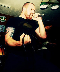 The image captures a live music performance, focusing on a bearded vocalist with a muscular build and tattoos on his arm. He is wearing a black t-shirt and is holding a microphone tightly while singing or screaming into it with intense emotion and energy. His facial expression and posture suggest that he is performing in a high-energy rock or metal concert. The dimly lit background includes a guitarist playing and a bar or club setting, with visible wall decorations, including a Brooklyn Brewery logo. The lighting, color grading, and grainy texture of the image add a raw, underground feel, often associated with hardcore, punk, or metal gigs in small venues. The coiled microphone cable in his hand and his aggressive stance further emphasize the powerful stage presence and passionate performance. This image likely showcases an artist fully immersed in their music, engaging with the audience through a high-impact performance.