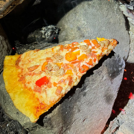 Slice of pizza with red peppers and sausage on a stone near a campfire. Brightly lit, contrasting with dark, smoky background.