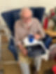 Elderly man with white beard sits in a blue chair, holding a blue mug and gift set