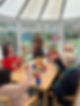 A woman guides elderly people in a crafting activity around a wooden table in a bright room