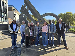 A group of six people smiling in front of a large, abstract metal sculpture under a clear blue sky
