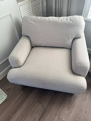 A plush, light gray armchair positioned in a cozy corner with light wood flooring and soft, neutral drapes