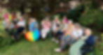 A group of elderly people sits outdoors, smiling and holding rainbow flags and wearing leis