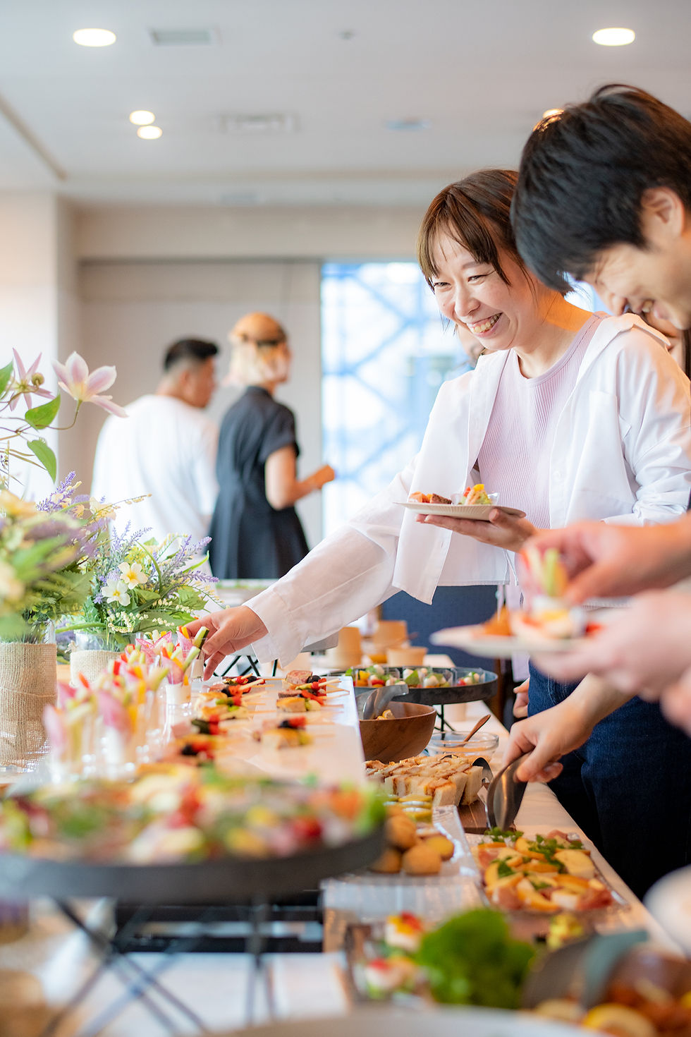 室内のパーティー会場で、笑顔の男女が色とりどりのフィンガーフードを取り分けるケータリングの様子。