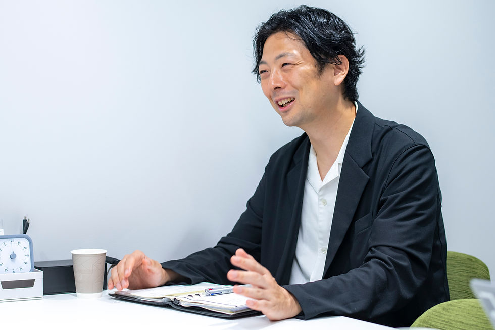 机の前で笑顔で話すボンディッシュ株式会社代表取締役社長の上形秀一郎。ノートやペン、紙コップ、時計が置かれた明るいオフィス風の室内。