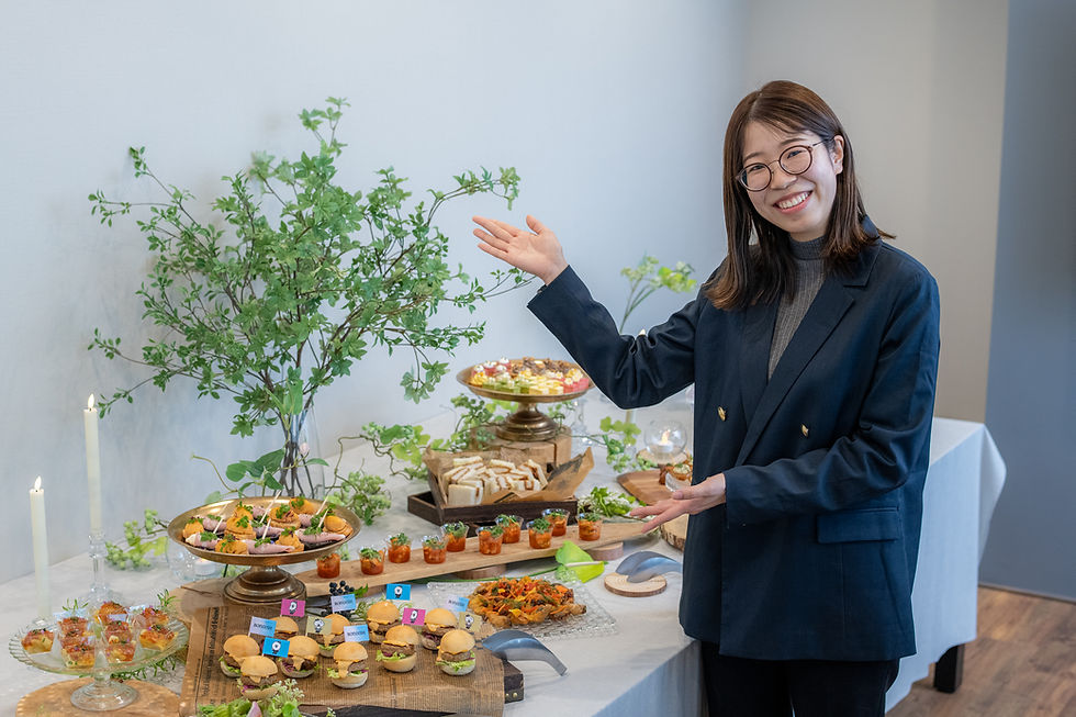 笑顔の女性がネイビーのスーツで料理ビュッフェを紹介。緑の枝やキャンドル、ミニバーガーに企業ロゴの旗が立っている。
