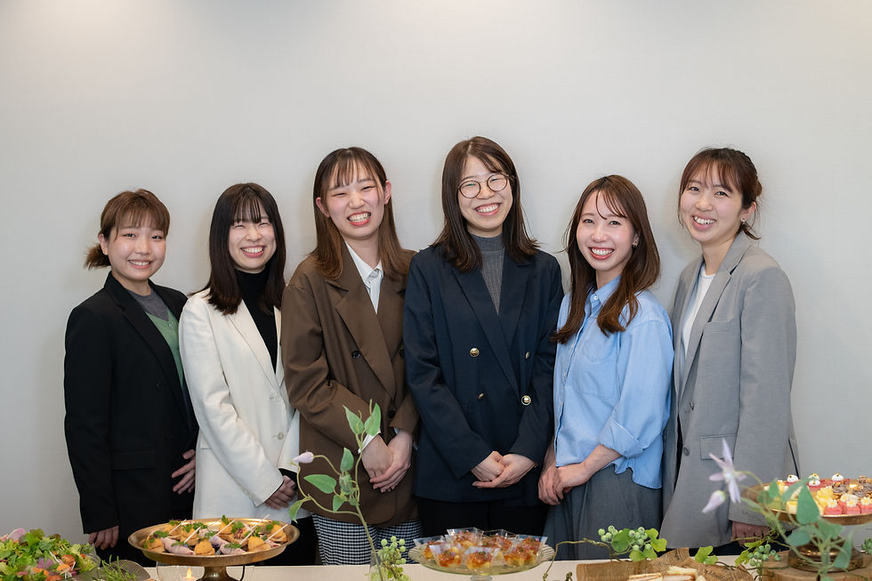 ビジネス服の笑顔の女性6人が白い壁の前に並び、手前のビュッフェ料理を囲んでいる。ケータリングのプランナーおよびコーディネーター。