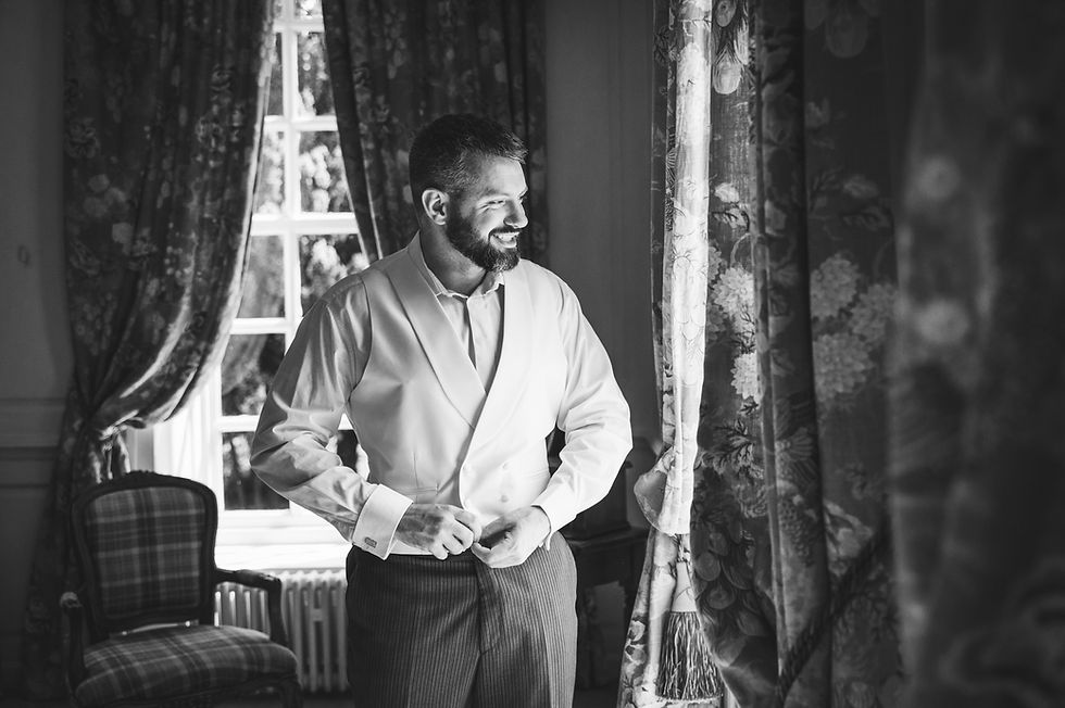 Gosfield Hall Wedding Photographer captures the groom getting ready in the elegant suite at Gosfield Hall, smiling as he buttons his waistcoat before the ceremony.