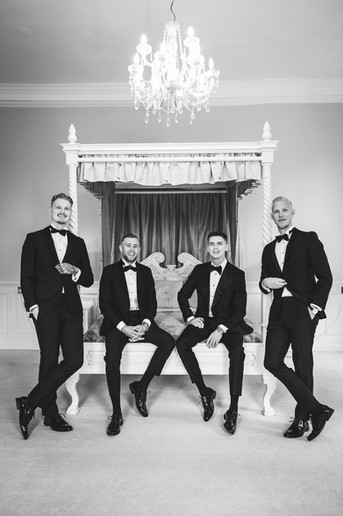 Gosfield Hall Wedding Photographer capturing the groom and his groomsmen dressed in tuxedos, sharing a relaxed moment together in the elegant Queen’s Room.