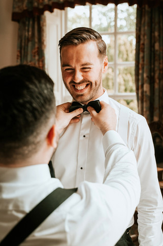A candid moment of the groom laughing as his best man adjusts his bow tie before the ceremony.