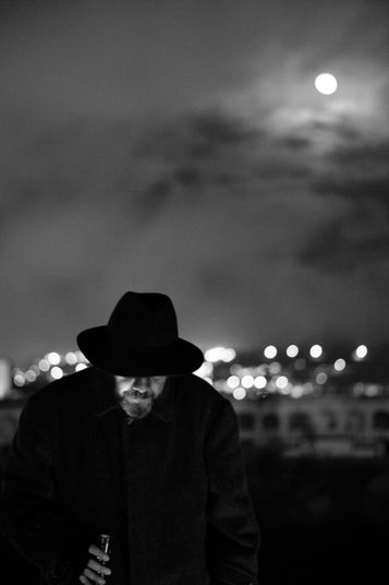 Hombre con abrigo y sombrero retratado de noche en el Cementerio del Tejar de Quito, en una escena en blanco y negro de alto contraste con luces desenfocadas y la luna en composición diagonal