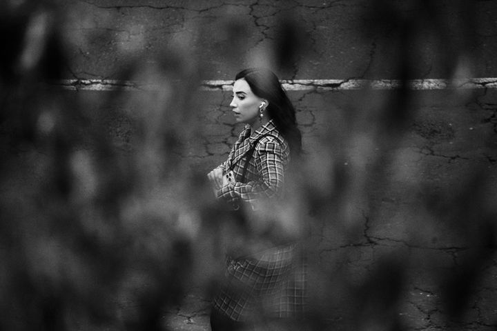 Mujer caminando en la calle en fotografía en blanco y negro, capturada con teleobjetivo y enmarcada por vegetación desenfocada en primer plano.