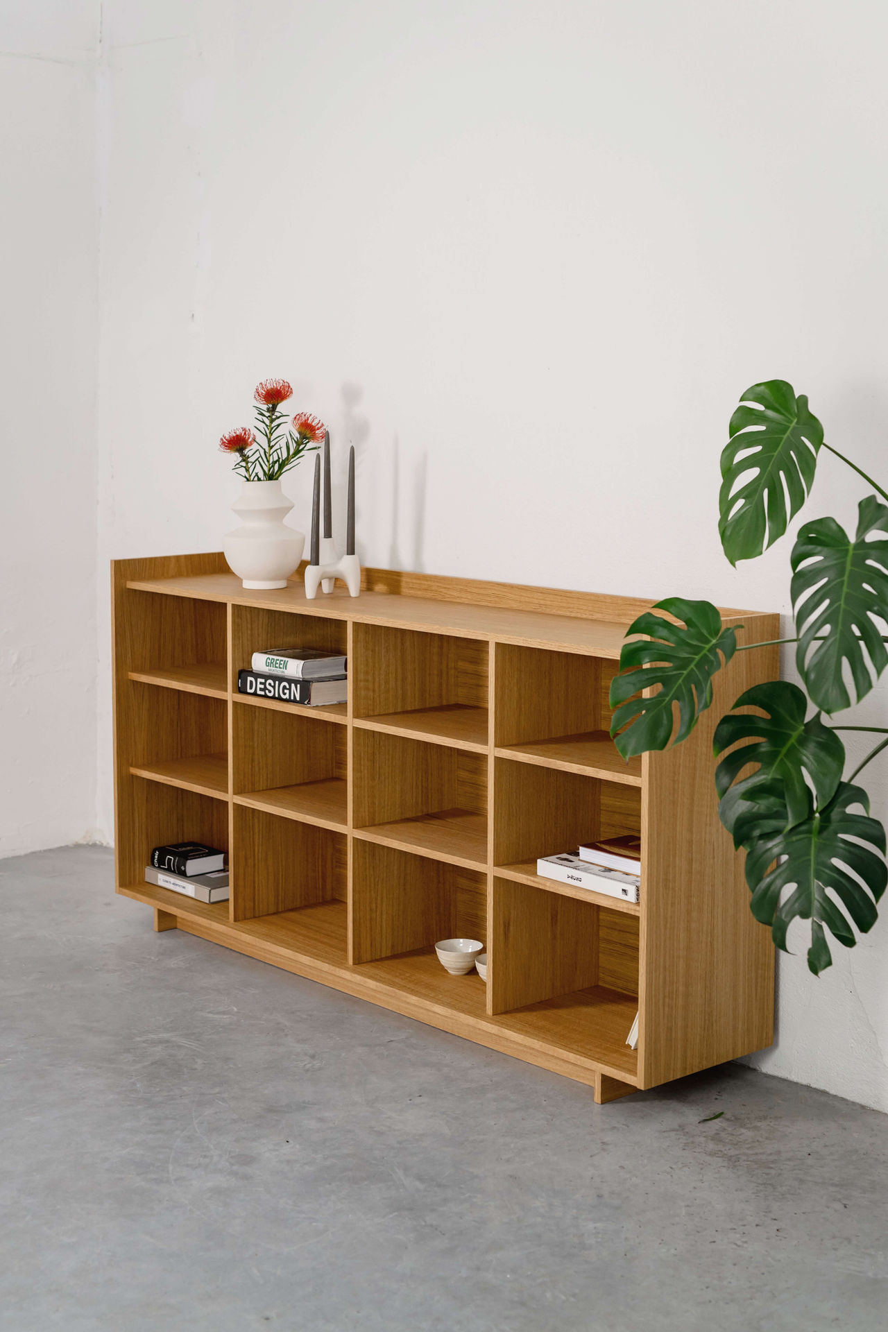 Wooden shelving unit with books, a vase of flowers, and candles on top, and a large plant next to it