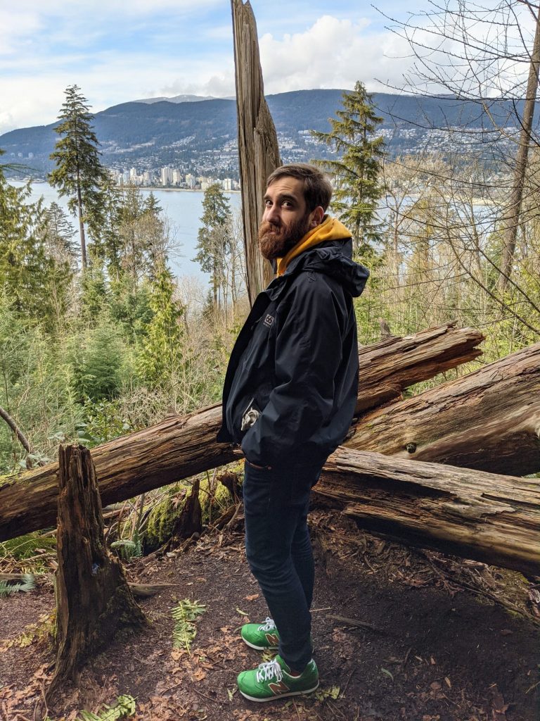 A bearded man stands on a log by a serene lake, surrounded by trees and reflecting water.
