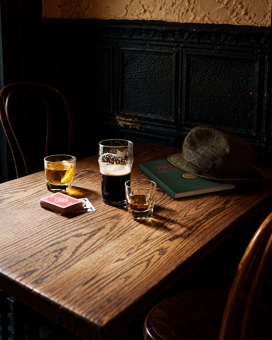 A pub table on a Sunday afternoon: a pint of stout, two whiskies, a deck of cards and a tweed cap resting on a copy of the Roman Sundays guide