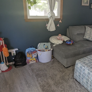 Living room with gray sectional, ottoman, and designated toddler play corner with baskets and toys.
