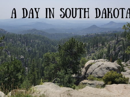 An image overlooking the forests and rolling Black Hills. Wildfire smoke covers the distant hills on a sunny day. Black text says A Day in South Dakota in handwritten font.