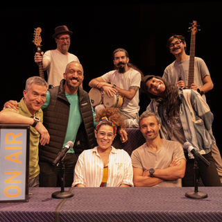 eight diverse, smiling people sit and stand together around a table. Three of them have musical instruments
