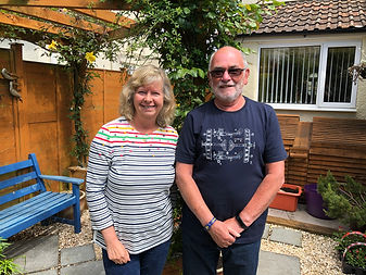 A husband and wife pet and house sitting team, living in Somerset, in their garden, underneath a pergola with climbing plants