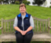 A female pet sitter, living in Dorset specialising cats sitting on a wooden bench wearing a navy body warmer with a green slope and trees behind