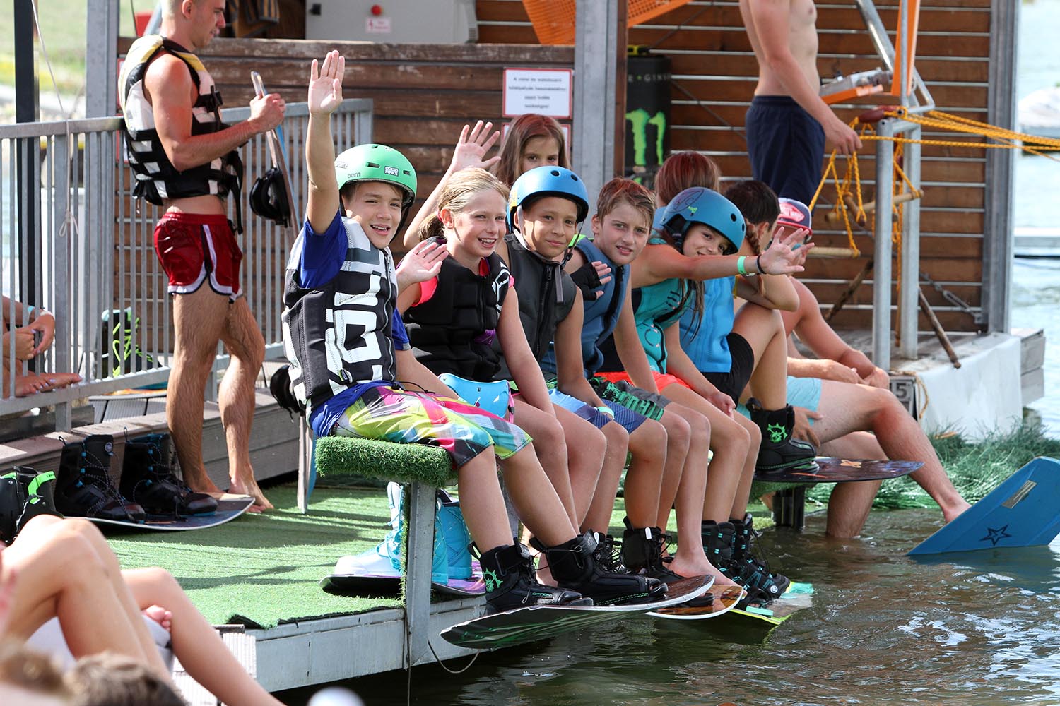 Group of kids with helmets and wakeboards sitting on the dock at Central Wakeboard Park, cable tower and lake behind them