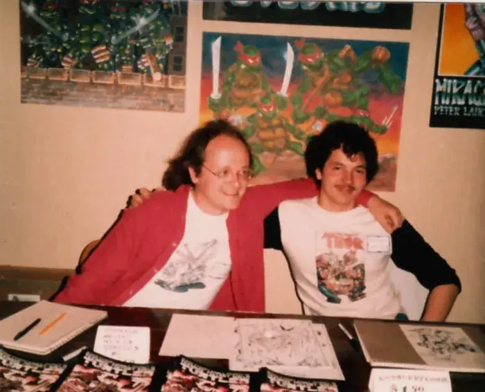 Kevin Eastman and Peter Laird sit side-by-side at a convention table with their arms around each other, smiling in front of Teenage Mutant Ninja Turtles posters and artwork, with TMNT comic issues and sketch pages spread out on the table.