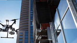 A drone hovers near a modern skyscraper reflecting sunlight. The scene is bright with a blue sky background and tall glass windows.