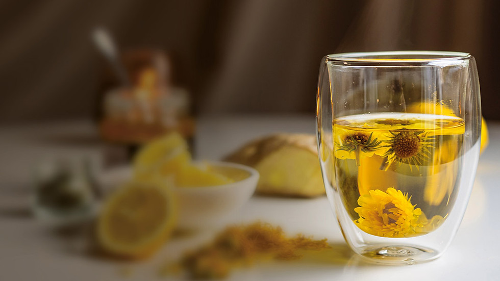Auf einem Tisch steht ein Teeglas mit frischem Tee, der gelbe Blüten enthält. Im Hintergrund Zitrone, Ingwer, Gewürze, Honig