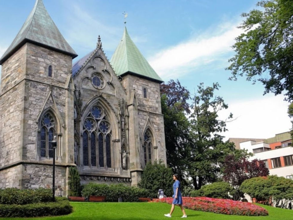 Stavanger Cathedral in Stavanger, Norway, a grey stone church with green copper roofs and Gothic windows, seen from a park lawn with flowers and trees in the foreground