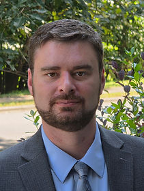 Author, Timothy J. McCollum II, wearing a black suit and blue tie. The background is an apple tree and various other foliage.