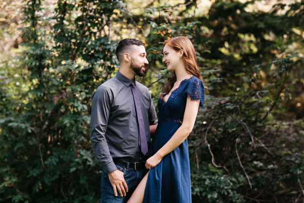 A man and woman in formal attire smiling at each other in front of large green bushes.