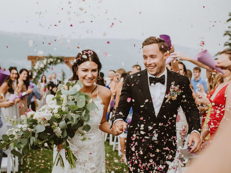 A newlywed man and woman laughing as they walk hand-in-hand down their outdoor ceremony aisle, while guests toss colourful confetti around them.